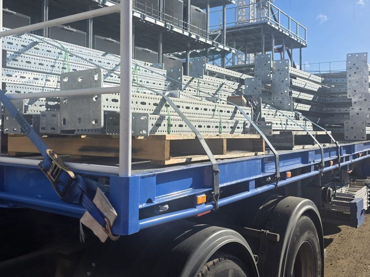 Structural steel loaded on a TLH trailer beside a building under construction, with a tower crane overhead.